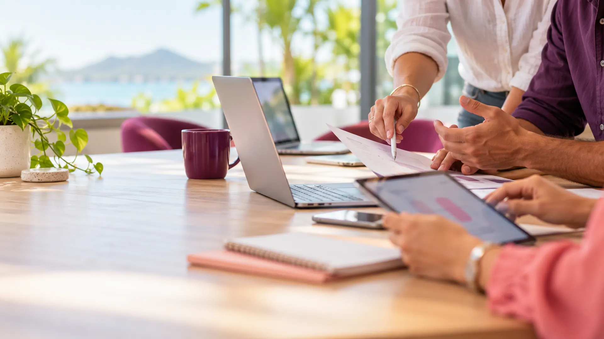 Hands around a meeting table sharing documents on laptops and a tablet
